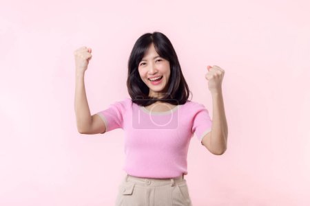 Téléchargez les photos : Portrait jeune belle femme asiatique sourire heureux avec poing vers le haut expression geste de victoire joyeuse sa réussite sur fond de studio pastel rose. Journée femme gagnant concept de célébration. - en image libre de droit