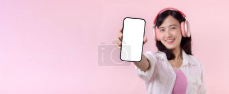 Foto de Feliz sonriente mujer asiática alegre con auriculares inalámbricos que muestran pantalla en blanco teléfono móvil o nuevo teléfono inteligente aplicación de música mockup anuncio aislado en rosa fondo del estudio. - Imagen libre de derechos