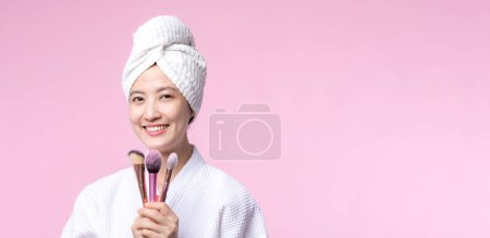 Téléchargez les photos : Portrait jeune femme asiatique heureuse avec naturel maquillage visage tenant peau cosmétique poudre rougisseur isolé sur fond rose. Femme appliquer un traitement de brosse de soins de la peau. produit de beauté, concept de cosmétologie. - en image libre de droit
