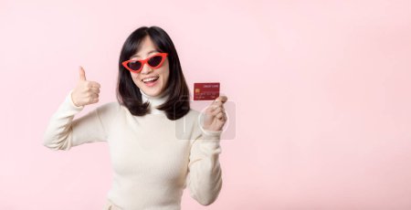 Téléchargez les photos : Fascinant joyeux amusant jeune femme d'origine asiatique 20 ans avec porter des lunettes de soleil porte chemise blanche tenir dans la main carte bancaire isolé sur pastel clair rose fond studio portrait. - en image libre de droit