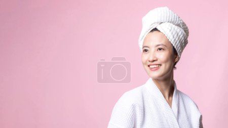 Foto de Hermosa mujer asiática joven feliz con la piel limpia de la cara en toalla y albornoz, traje de spa sobre fondo rosa. Cuidado de la piel, tratamiento, terapia de bienestar, cuidado facial, salud femenina de belleza, concepto de cosmetología - Imagen libre de derechos
