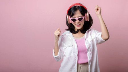 Téléchargez les photos : Jeune modèle de femme asiatique heureuse avec des lunettes de soleil tendance élégantes profiter de l'écoute de la musique par casque audio et la danse isolé sur fond de studio rose. technologie, girl fashion, concept accessoire. - en image libre de droit