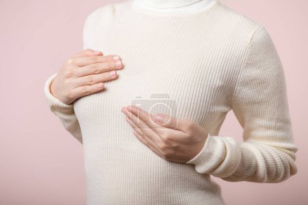 Téléchargez les photos : Les mains de la femme vérifient des morceaux sur son sein pour détecter des signes de cancer du sein sur fond rose. Concept de soins de santé. - en image libre de droit