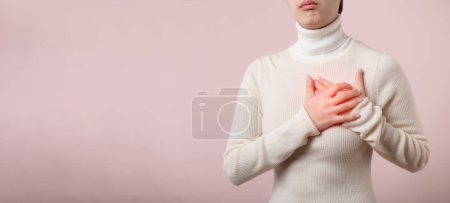 Foto de Joven mujer asiática que sufre de un ataque al corazón en el fondo del estudio rosa claro. Calambres dolorosos, Enfermedades del corazón, Presionar el pecho con expresión dolorosa. Concepto sanitario. - Imagen libre de derechos