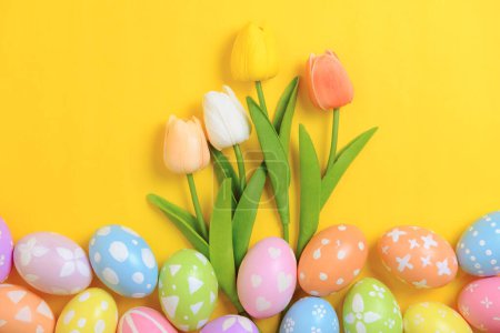 Happy Easter holiday greeting card concept. Colorful Easter Eggs and spring flowers on yellow background. Top view, flat lay, copy space.