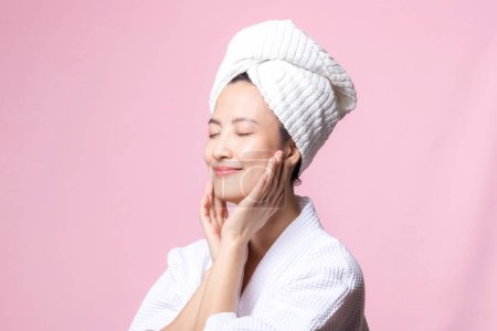 Téléchargez les photos : Belle jeune femme asiatique heureuse avec peau de visage propre en serviette et peignoir, costume de spa sur fond rose. Soins de la peau, traitement, thérapie de bien-être, soins du visage, beauté santé féminine, concept de cosmétologie - en image libre de droit