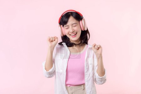 Foto de Sonrisa asiática bonita modelo persona escuchar música canción y disfrutar de la danza con auriculares inalámbricos en línea sonido de radio de audio. Diversión positiva salió alegre joven mujer femenina en rosa aislado estudio de fondo - Imagen libre de derechos