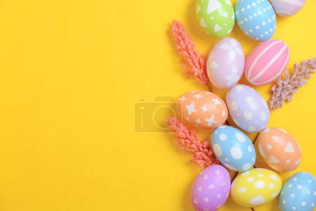 Happy Easter holiday greeting card concept. Colorful Easter Eggs and spring flowers on yellow background. Top view, flat lay, copy space.