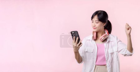 Foto de Retrato atractiva mujer asiática joven sonrisa feliz usando teléfono inteligente con auriculares, auriculares aislados en el fondo del estudio rosa. Persona bastante femenina usando el teléfono móvil. Música concepto de estilo de vida en línea. - Imagen libre de derechos
