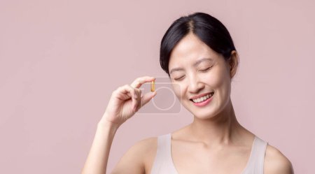 Téléchargez les photos : Jeune portrait asiatique femme sourire heureux visage avec vitamine pilule de nutrition. Jolie fille mignonne femme tenant capsule santé supplément soin de la peau isolé sur fond rose. Concept de médicament. - en image libre de droit