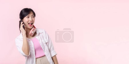 Téléchargez les photos : Portrait attrayant jeune femme asiatique sourire heureux en utilisant smartphone avec écouteur, casque isolé sur fond de studio rose. Jolie femme utilisant un téléphone portable. Musique concept de style de vie en ligne. - en image libre de droit