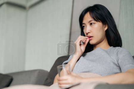 Téléchargez les photos : Asiatique jeune femme prendre un médicament pilule tandis que la pose sur canapé - en image libre de droit