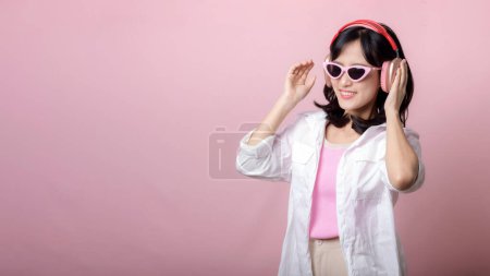 Foto de Joven feliz mujer asiática modelo con gafas de sol de moda con estilo disfrutar de escuchar música por auriculares de audio y baile aislado en el fondo del estudio rosa. tecnología, moda de chica, concepto de accesorio. - Imagen libre de derechos