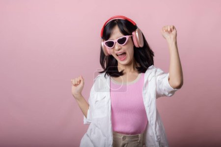 Téléchargez les photos : Jeune modèle de femme asiatique heureuse avec des lunettes de soleil tendance élégantes profiter de l'écoute de la musique par casque audio et la danse isolé sur fond de studio rose. technologie, girl fashion, concept accessoire. - en image libre de droit
