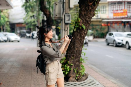 Téléchargez les photos : Jeune voyageur asiatique sac à dos femme en utilisant un appareil photo numérique compact, profiter de la rue lieu culturel local et sourire. - en image libre de droit
