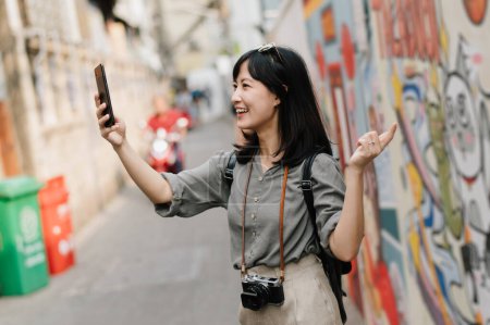 Foto de Young Asian woman backpack traveler using mobile phone, enjoying street cultural local art wall. - Imagen libre de derechos