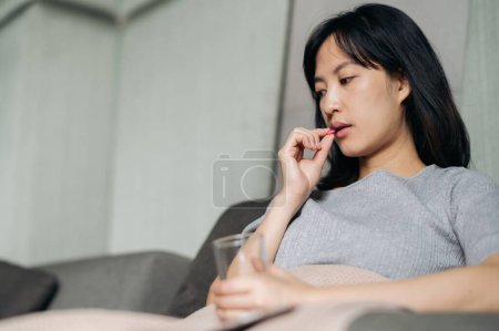 Téléchargez les photos : Asiatique jeune femme prendre un médicament pilule tandis que la pose sur canapé - en image libre de droit