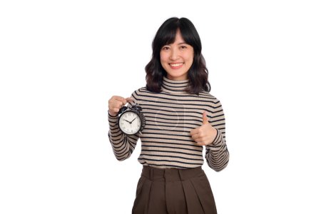 Foto de Sonriente alegre atractiva joven morena asiática mujer vistiendo suéter camisa de pie celebrar reloj mostrando el pulgar hacia arriba mirando cámara sobre fondo blanco, estudio retrato. - Imagen libre de derechos