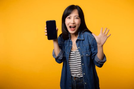 Photo for Portrait beautiful asian young woman happy smile dressed in denim jacket showing smartphone screen isolate on yellow studio background. New smartphone application concept - Royalty Free Image