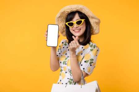 Téléchargez les photos : Portrait heureuse jeune femme asiatique montrant smartphone écran vierge avec robe de printemps à la mode, chapeau, lunettes de soleil mode et sac en papier isolé sur fond jaune. Vente d'été concept de shopping. - en image libre de droit