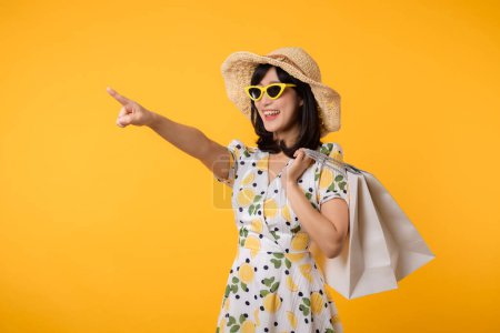 Téléchargez les photos : Portrait attrayant jeune femme asiatique heureuse avec robe de printemps à la mode, chapeau, lunettes de soleil mode et sac en papier isolé sur fond jaune. Vente d'été concept de shopping. - en image libre de droit