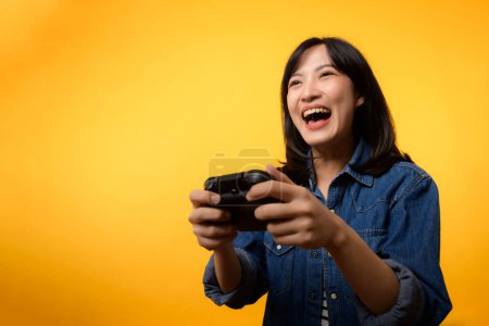 Téléchargez les photos : Portrait asiatique jeune femme avec un sourire heureux succès portant des vêtements en denim tenant manette de commande et de jouer à un jeu vidéo. Fun and relax hobby divertissement style de vie concept. - en image libre de droit