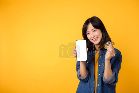 Foto de Feliz asiática joven mujer con camiseta amarilla camisa de mezclilla que sostiene moneda cripto digital moneda y teléfono inteligente aislado en el fondo amarillo. Concepto financiero de moneda digital. - Imagen libre de derechos