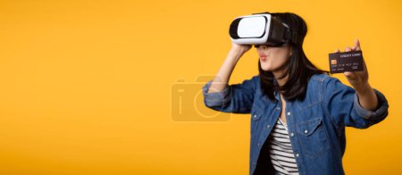 Téléchargez les photos : Asiatique jeune femme heureux succès portant réalité virtuelle jeu de casque divertissement et tenant carte de crédit isolé sur fond jaune. Gagnant du concept de cyberfutur jeu vidéo. - en image libre de droit