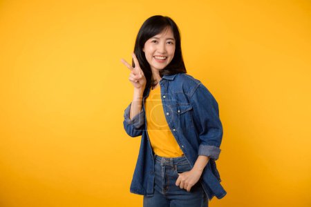 Téléchargez les photos : Jeune souriante joyeuse femme amicale d'origine asiatique porter chemise en jean t-shirt jaune montrant signe de victoire isolé sur fond jaune portrait studio. Concept de style de vie. - en image libre de droit