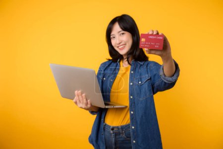 Téléchargez les photos : Jeune femme heureuse d'origine asiatique porter t-shirt jaune chemise en denim en utilisant ordinateur portable pc détenir carte bancaire de crédit achats livraison de commande en ligne isolé sur fond jaune uni. - en image libre de droit