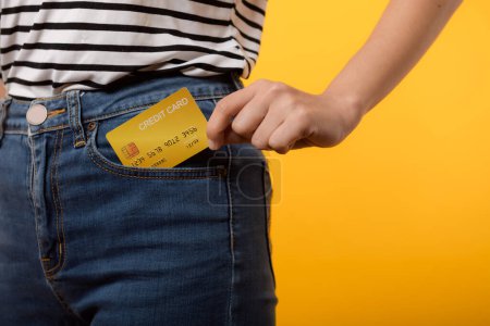 Téléchargez les photos : Main de la personne femme tenant la carte de crédit en plastique de la poche plantes de jean bleu isolé sur fond jaune studio. achat en ligne paiement, devise, facture, argent de paiement, concept de finance. - en image libre de droit