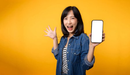 Téléchargez les photos : Portrait belle asiatique jeune femme sourire heureux vêtu d'une veste en denim montrant écran smartphone isoler sur fond jaune studio. Nouveau concept d'application smartphone - en image libre de droit