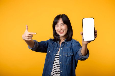 Téléchargez les photos : Portrait belle asiatique jeune femme heureux sourire habillé en denim veste montrant écran smartphone avec pointant doigt geste de la main pour téléphone mobile isoler sur fond jaune studio. - en image libre de droit