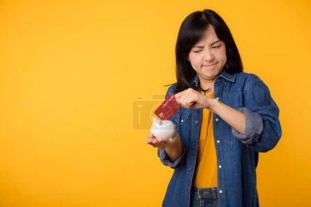 Téléchargez les photos : Malheureuse jeune femme asiatique portant t-shirt jaune chemise en denim tirant carte de crédit de tirelire isolé sur fond jaune. Paiement dette concept financier. - en image libre de droit