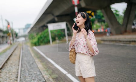Photo for Young Asian woman traveler with weaving basket using a mobile phone beside railway train station in Bangkok. Journey trip lifestyle, world travel explorer or Asia summer tourism concept. - Royalty Free Image