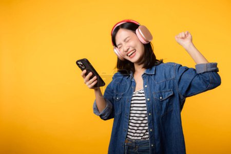 Téléchargez les photos : Portrait jeune femme asiatique gaie profiter de l'écoute audio par application de musique smartphone contre jaune. Heureuse femme souriante avec casque. Son, loisirs, mode de vie, concept technologique - en image libre de droit