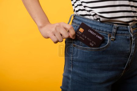 Téléchargez les photos : Main de la personne femme tenant la carte de crédit en plastique de la poche plantes de jean bleu isolé sur fond jaune studio. achat en ligne paiement, devise, facture, argent de paiement, concept de finance. - en image libre de droit