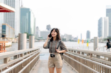 Téléchargez les photos : Young Asian woman backpack traveler using mobile phone in express boat pier on Chao Phraya River in Bangkok. Traveler checking out side streets. - en image libre de droit