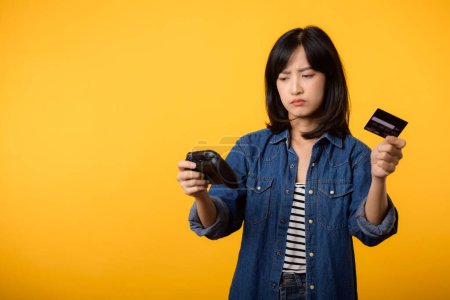 Photo for Portrait asian young woman with doubt face wearing denim clothes holding joystick controller and credit card. Shopping online game entertainment lifestyle concept. - Royalty Free Image