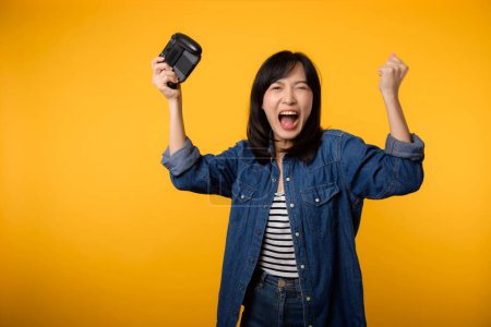Foto de Retrato de mujer joven asiática con feliz éxito sonrisa usando ropa de mezclilla sosteniendo controlador de joystick y jugando videojuego. Diversión y relax hobby entretenimiento concepto de estilo de vida. - Imagen libre de derechos