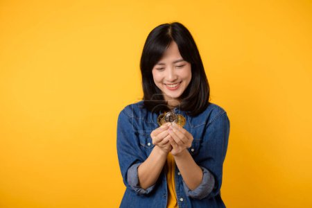 Photo for Happy asian young woman wearing yellow t-shirt denim shirt holding digital crypto currency coin isolated on yellow background. Digital currency financial concept. - Royalty Free Image