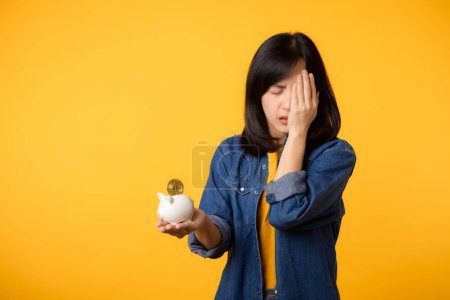 Photo for Unhappy young asian woman wearing yellow t-shirt denim shirt pulling digital coin crypto currency out of piggy bank isolated on yellow background. Payment digital money debt financial concept. - Royalty Free Image