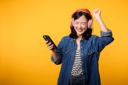 Téléchargez les photos : Portrait jeune femme asiatique gaie profiter de l'écoute audio par application de musique smartphone contre jaune. Heureuse femme souriante avec casque. Son, loisirs, mode de vie, concept technologique - en image libre de droit