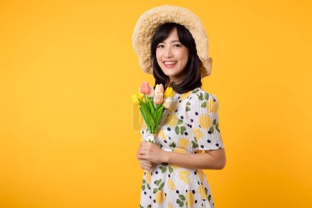 Foto de Estudio retrato bastante joven mujer asiática feliz sonrisa vestirse moda primaveral con sombrero celebración tulipán ramo de flores sobre fondo amarillo estudio. Concepto de vacaciones persona. - Imagen libre de derechos