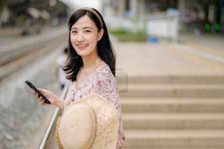 Photo for Young Asian woman traveler with weaving hat using a mobile phone beside railway train station in Bangkok. Journey trip lifestyle, world travel explorer or Asia summer tourism concept. - Royalty Free Image