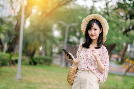 Photo for Portrait of young asian woman traveler with weaving hat and basket using mobile phone on green park nature background. Journey trip lifestyle, world travel explorer or Asia summer tourism concept. - Royalty Free Image