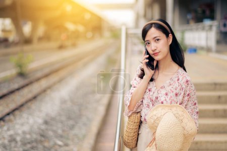 Foto de Joven viajera asiática con sombrero de tejer usando un teléfono móvil al lado de la estación de tren en Bangkok. Viaje viaje estilo de vida, viajero mundial o concepto de turismo de verano de Asia. - Imagen libre de derechos