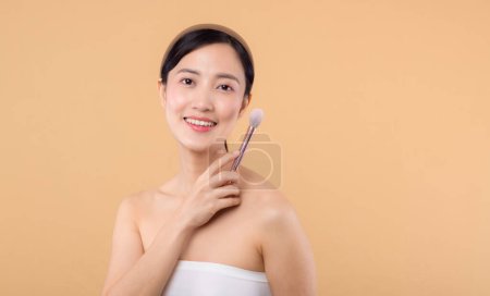 Téléchargez les photos : Portrait jeune femme asiatique heureuse avec naturel maquillage visage tenant peau cosmétique poudre rougisseur isolé sur fond beige. Femme appliquer un traitement de brosse de soins de la peau. produit de beauté, concept de cosmétologie - en image libre de droit