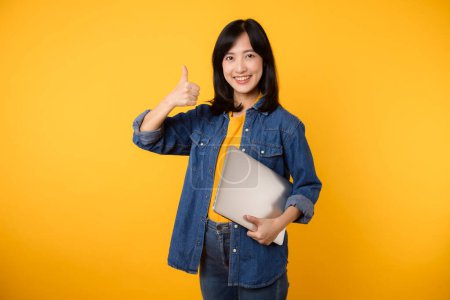 Téléchargez les photos : Portrait jeune femme asiatique portant un t-shirt jaune et une chemise en denim tenant un ordinateur portable isolé sur fond de studio jaune. Technologie ordinateur portable annonce concept. - en image libre de droit