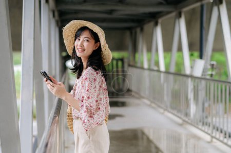 Photo for Asian young woman traveler with weaving basket using mobile phone and standing on overpass. Journey trip lifestyle, world travel explorer or Asia summer tourism concept. - Royalty Free Image